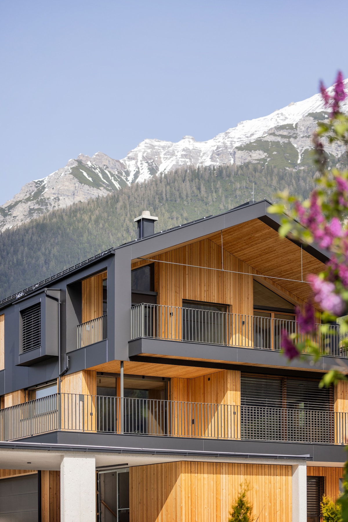 The modern building features a distinctive mix of wood and metal facades, emphasizing clean lines and geometric shapes. A backdrop of towering mountains is visible, with snow-capped peaks contrasting against the clear blue sky. Lush greenery is provided by nearby flowering plants.