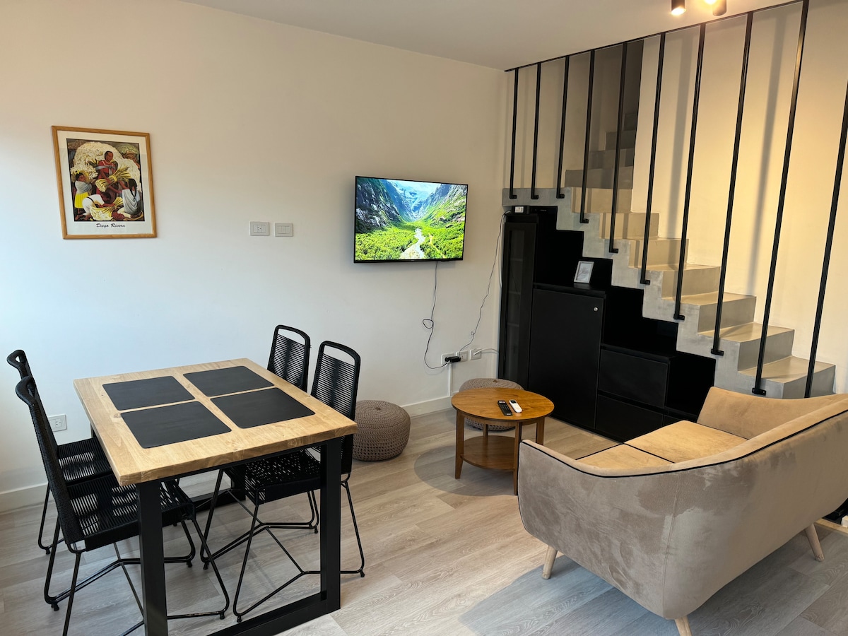 A modern living area is presented with a dining table set for six and a comfortable sofa. A flat-screen TV is mounted on the wall, showcasing scenic views. Stairs lead to the upper level, with a minimalist railing design enhancing the space's openness.