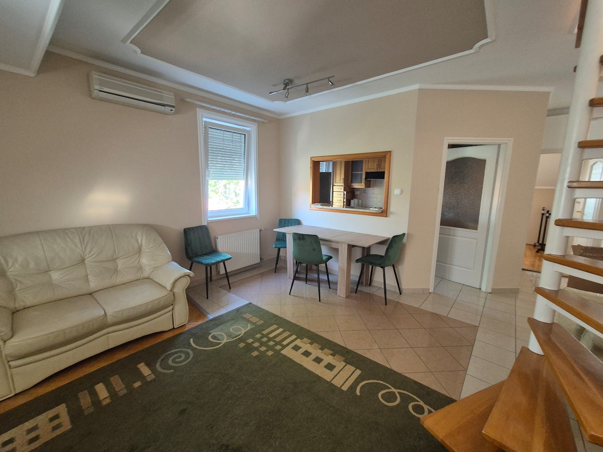 A light-filled living area features a comfortable cream sofa and a simple dining table surrounded by four chairs. The space includes a mirror that reflects the open kitchen area, with beige walls and tiled flooring enhancing the room's spacious feel.