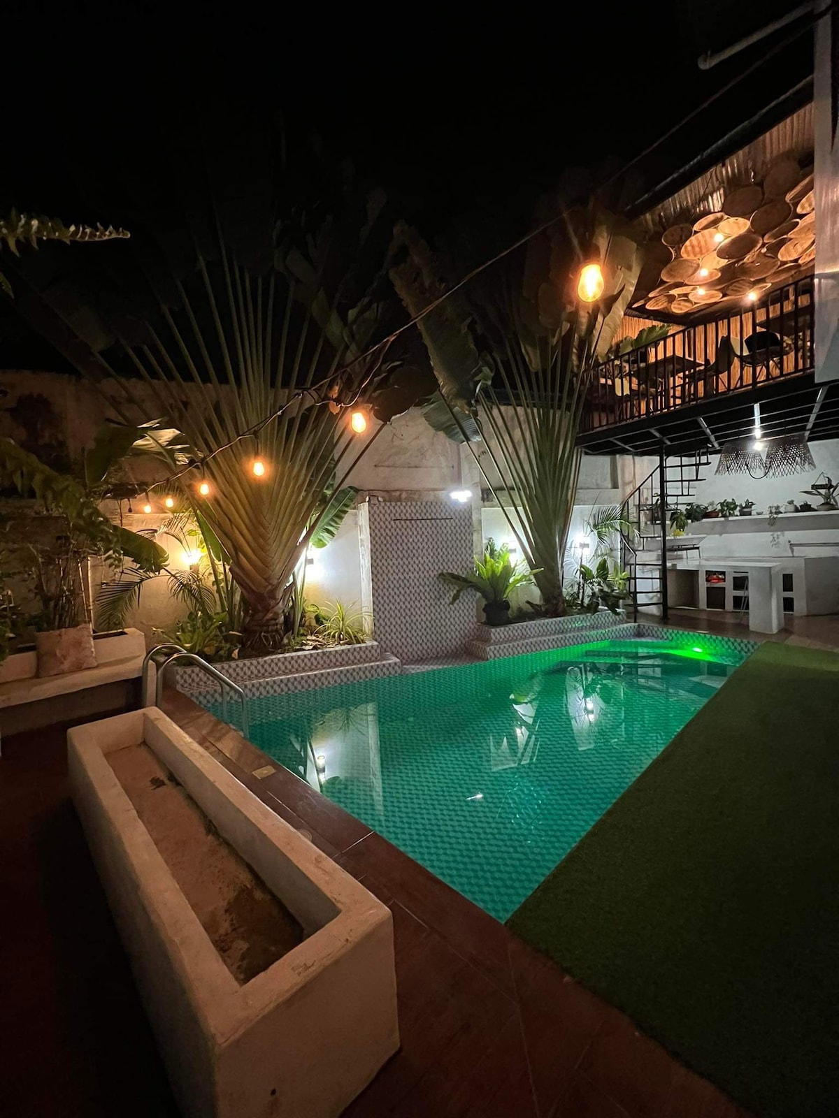 An illuminated private pool area is surrounded by lush greenery and tall palm trees. Soft lighting accentuates the space, creating a calming ambiance. The pool's turquoise water reflects the light, while a well-maintained area features a modern outdoor kitchen and vibrant plant arrangements nearby.