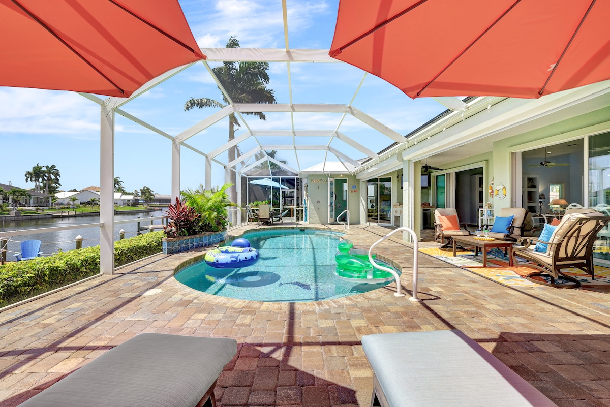 A landscaped outdoor area features a heated saltwater pool surrounded by loungers and colorful floats. Red umbrellas provide shade over seating areas. The screened enclosure offers views of the canal and neighboring homes, with lush greenery adding to the tranquil atmosphere.