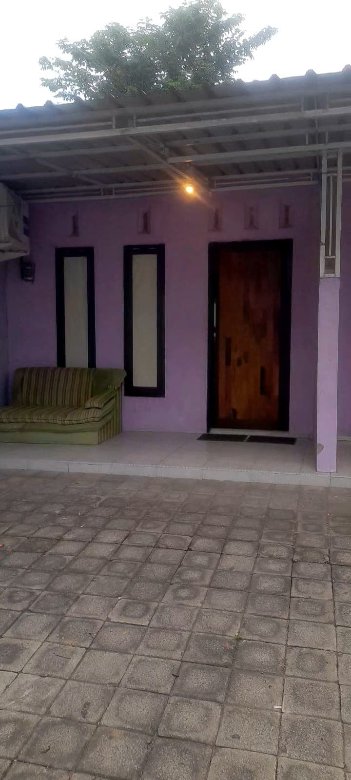 An entrance area is highlighted by a wooden door framed by narrow windows on either side. A green striped sofa is positioned against a light purple wall, providing a relaxed seating option. The paved flooring leads to an open space, creating a welcoming atmosphere.
