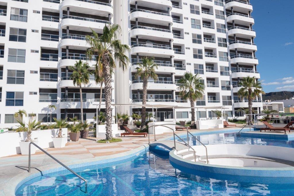 The image depicts a modern building featuring multiple levels with balconies and large windows. The foreground reveals a swimming pool with a jacuzzi area, surrounded by palm trees and lounge chairs, creating an inviting outdoor environment.