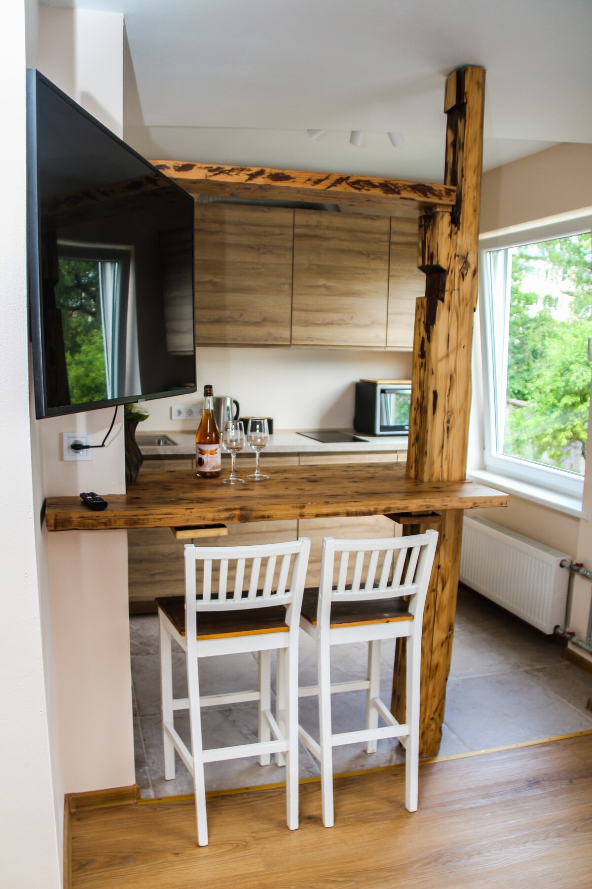 A compact kitchen area is highlighted by natural wood finishes, featuring a small bar with two white stools. A flat-screen TV is mounted on the wall, and glassware is arranged on the bar. Large windows allow natural light to illuminate the space.