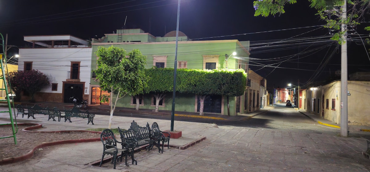 A quiet street scene is depicted at night, featuring charming benches arranged around a well-maintained park. Lush shrubs and trees provide greenery alongside the historic buildings. Streetlights illuminate the area, enhancing visibility and creating a peaceful atmosphere in this inviting neighborhood.