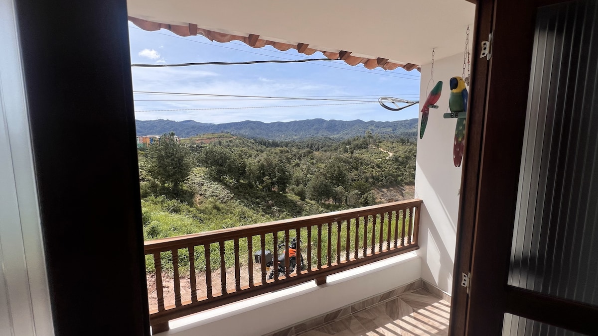 A private balcony features a wooden railing that overlooks a lush green landscape. Rolling hills and distant mountains create a natural backdrop, while a hint of wildlife decor adds a touch of charm to the outdoor space.