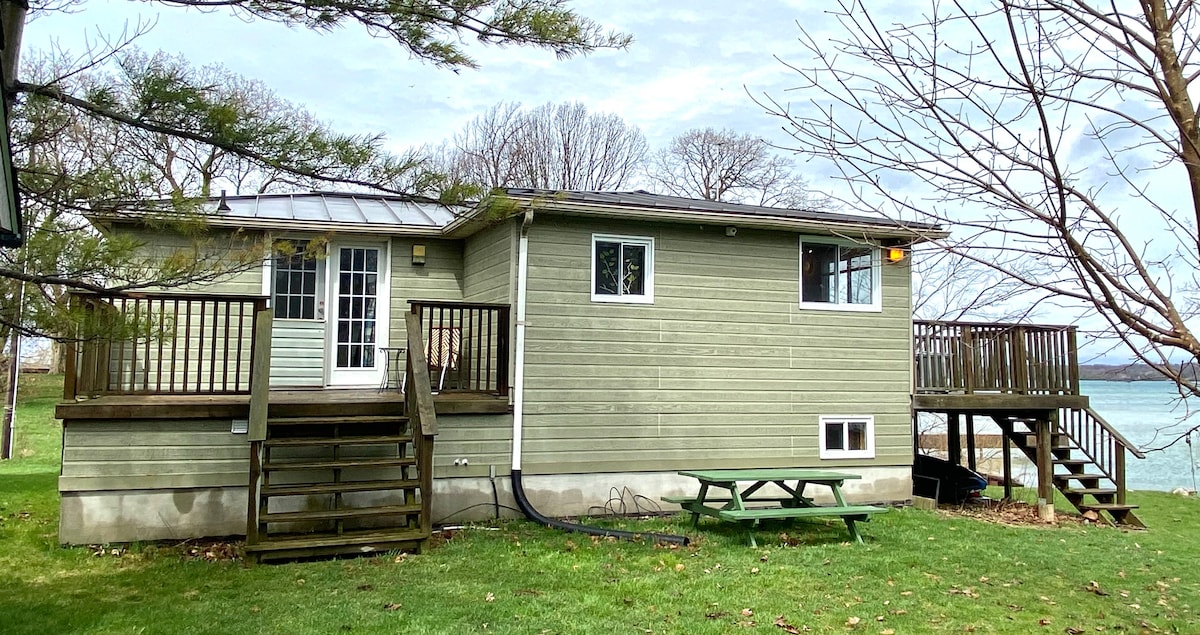 A wooden cottage is presented, featuring a light green exterior with a deck accessible by a set of stairs. A picnic table is situated on the grassy area in front. Large windows allow abundant natural light, while the backdrop includes bare trees and a view of the water.