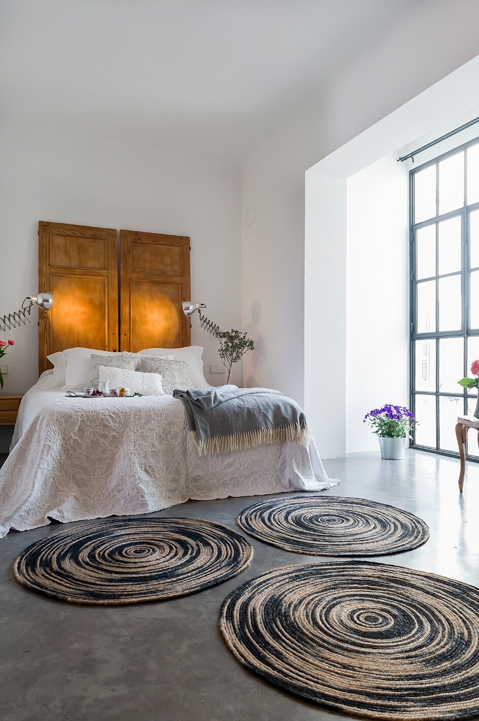 A spacious sleeping area features a double bed with a light-colored coverlet. Two warm bedside lights highlight wooden headboard elements. Below, circular patterned rugs contrast with the concrete flooring. A large window allows natural light to fill the room, with potted plants adding a touch of color.