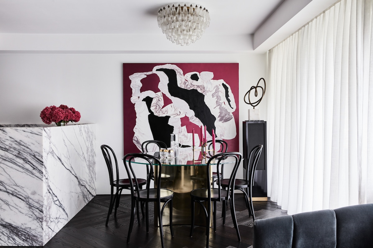 A stylish dining area features a round glass table surrounded by six black chairs. A marble counter serves as a stylish backdrop, complemented by a bold abstract wall art piece in shades of black and burgundy. Light filters through sheer curtains, enhancing the inviting atmosphere.