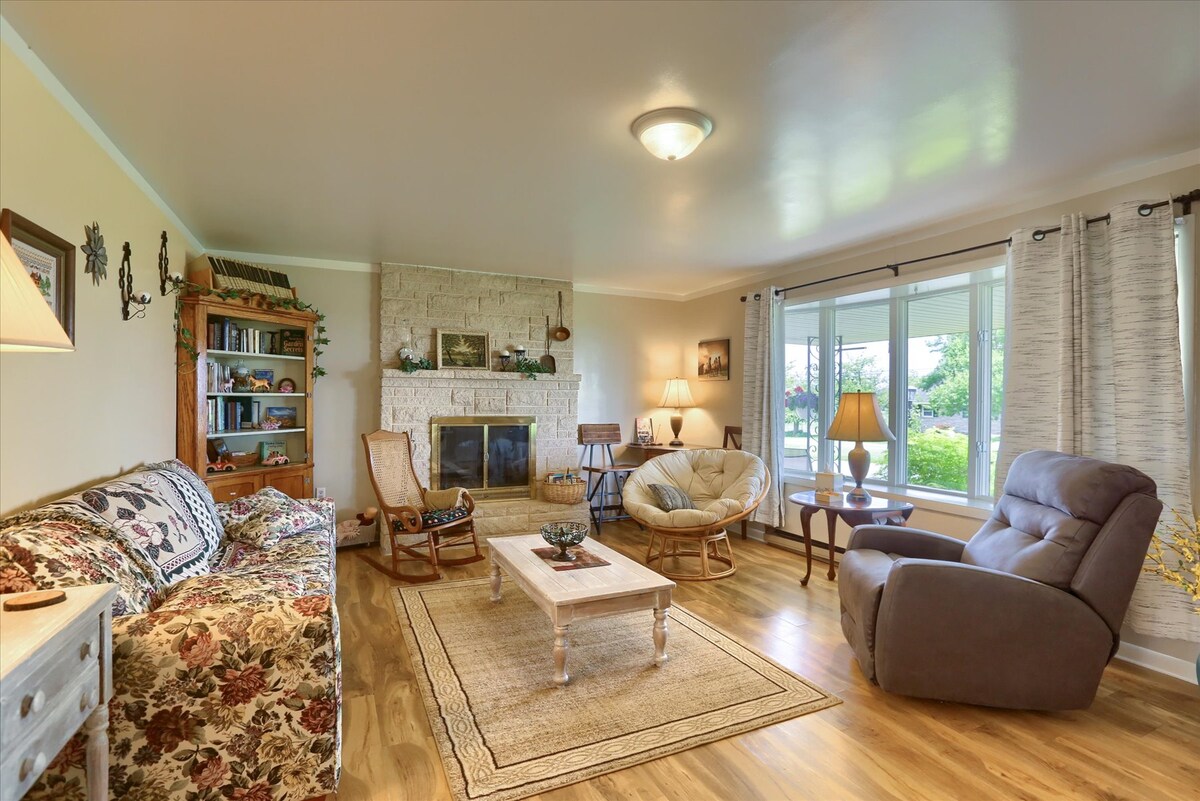 The living area features a mix of seating options, including a floral-patterned couch, rocking chairs, and a recliner. A wooden coffee table rests on a light area rug, while large windows allow natural light to fill the space. A stone fireplace is centrally located against a neutral wall.