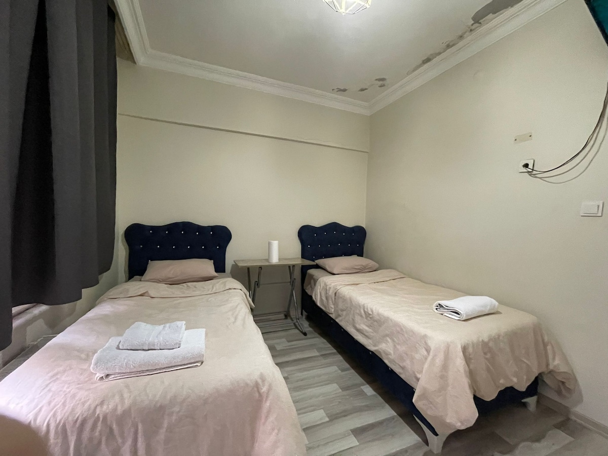A simple sleeping area features two beds with navy blue headboards, topped with light bedding and neatly folded towels. A small table is positioned between the beds, and natural light enters through a window, partially covered by dark curtains.