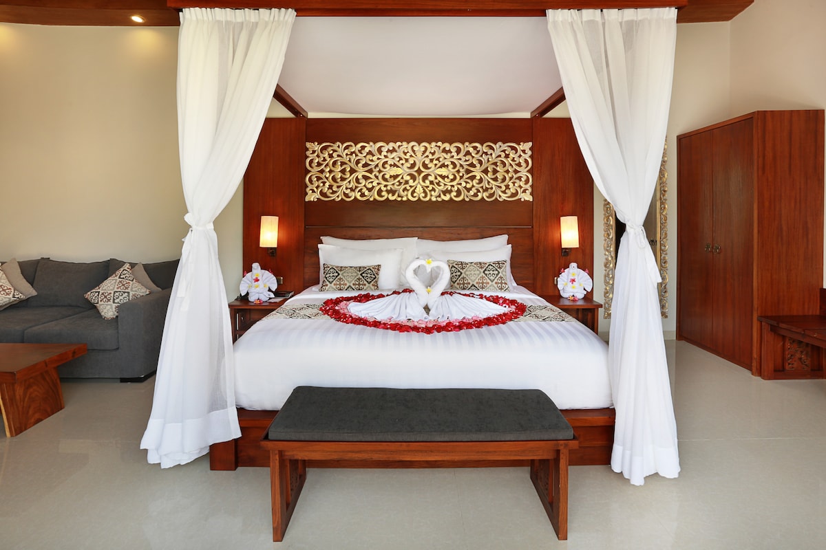 A spacious bedroom features a decorative wooden bed frame adorned with elegant carvings. Soft white drapes frame the bed, which is laid with petals and two swan-shaped towels. A comfortable seating area and wooden accents complete the inviting ambiance.