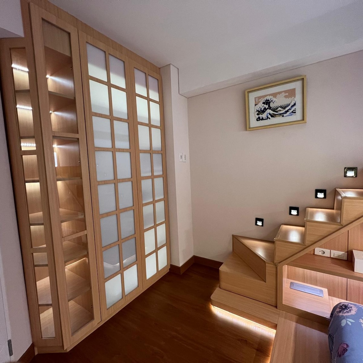 A modern wooden cabinet features glass doors, providing ample storage and a sleek appearance. A well-lit staircase with illuminated steps leads to the upper level, enhancing the aesthetic of the space. The warm wood tones contribute to a cohesive Japanese design.
