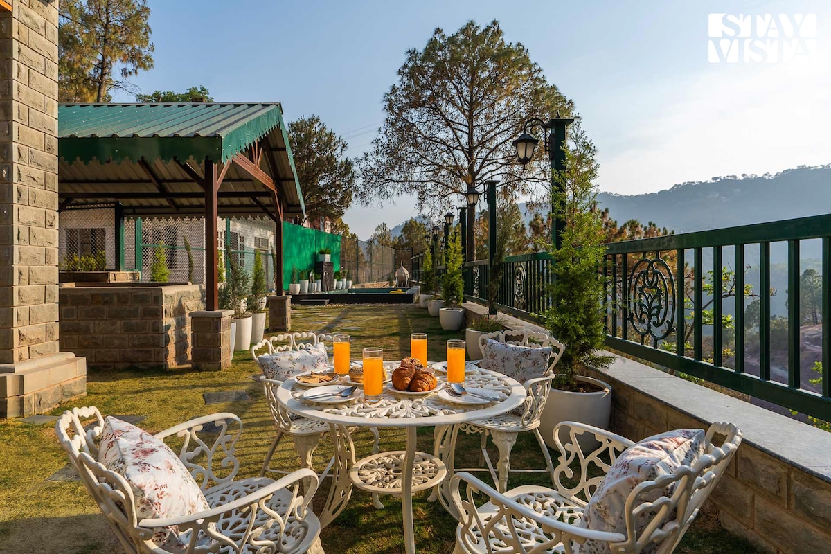 An outdoor seating area features a round table surrounded by four chairs, adorned with floral cushions. Bright orange drinks are placed on the table alongside pastries. Lush greenery and distant hills are visible, enhancing the serene atmosphere of the outdoor space.