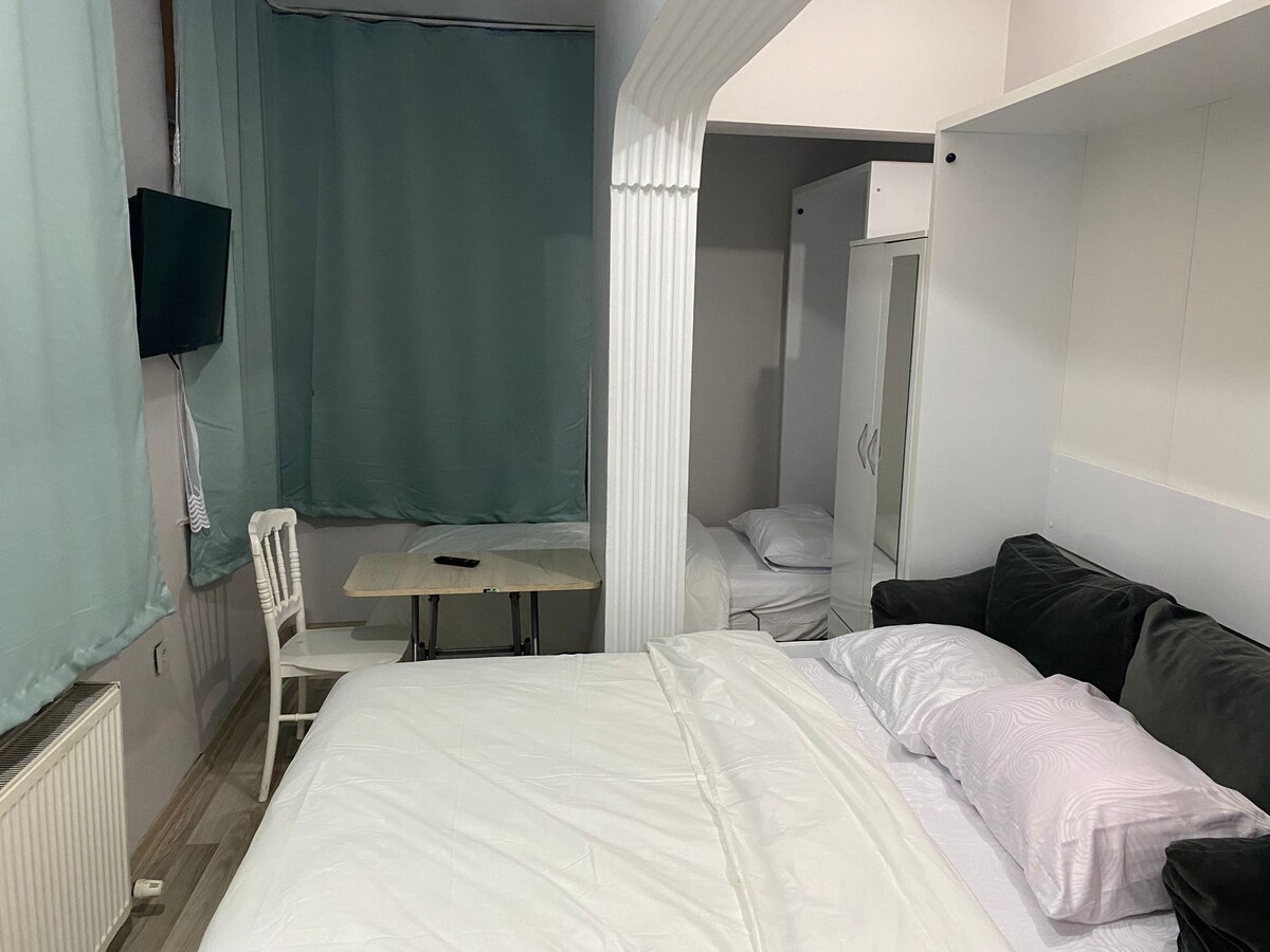 A cozy studio features a spacious bed with white linens, a small table with two chairs, and a separate sleeping area. Light green curtains cover the windows, while a television is mounted on the wall. A wardrobe provides additional storage options.