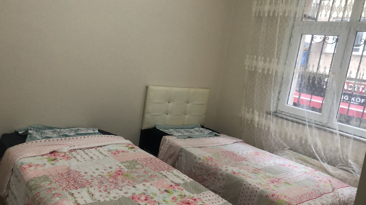 The room features two single beds, each adorned with floral-patterned bedding. A neutral-colored headboard complements the decor. Natural light filters through a lace curtain, illuminating the space. A large window offers a view of the surrounding area.