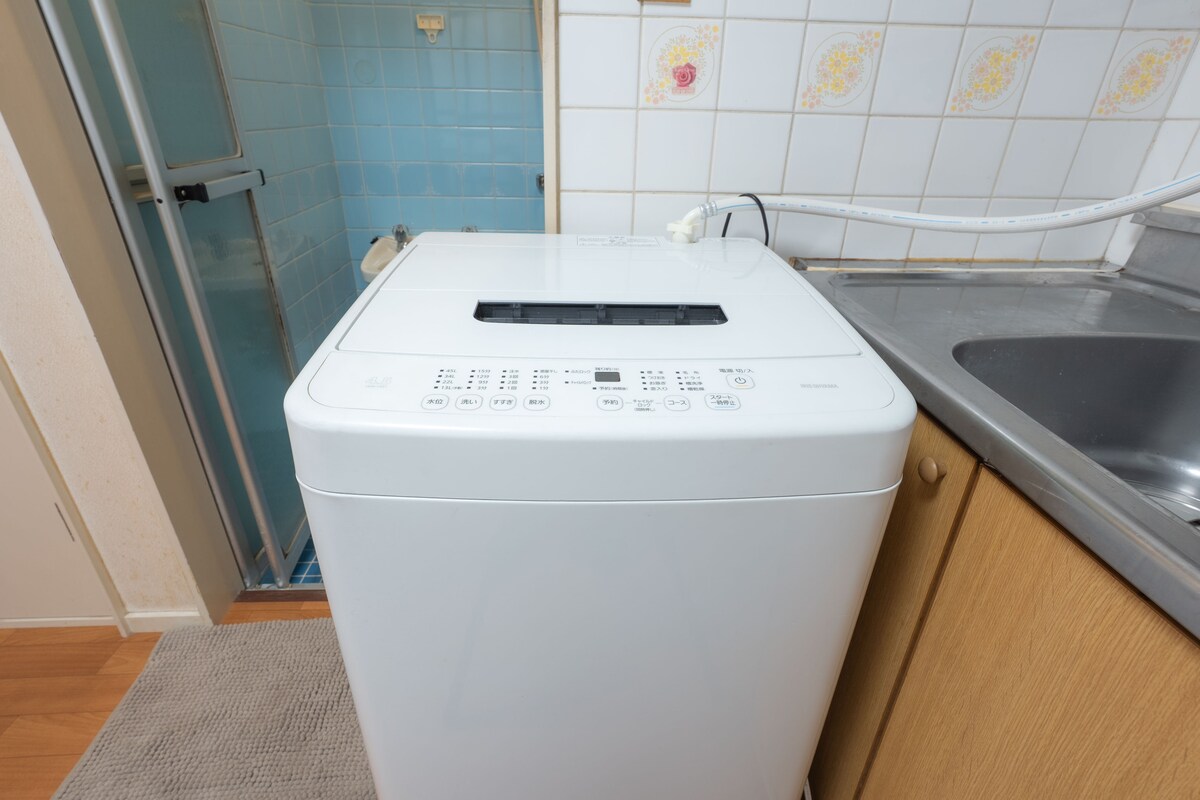A compact washing machine is positioned next to a sink and in close proximity to a bathroom area. The machine features a simple control panel with various buttons. Light wooden cabinetry complements the neutral tones of the flooring and surrounding walls.