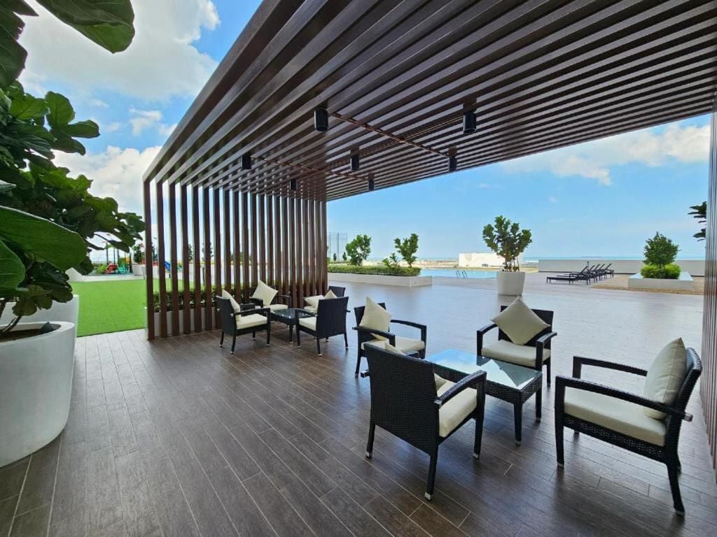 An outdoor lounge area features several seating arrangements with comfortable chairs and a glass table, surrounded by greenery. A shaded structure provides protection from the sun, while a view of the swimming pool and distant ocean is visible in the background.