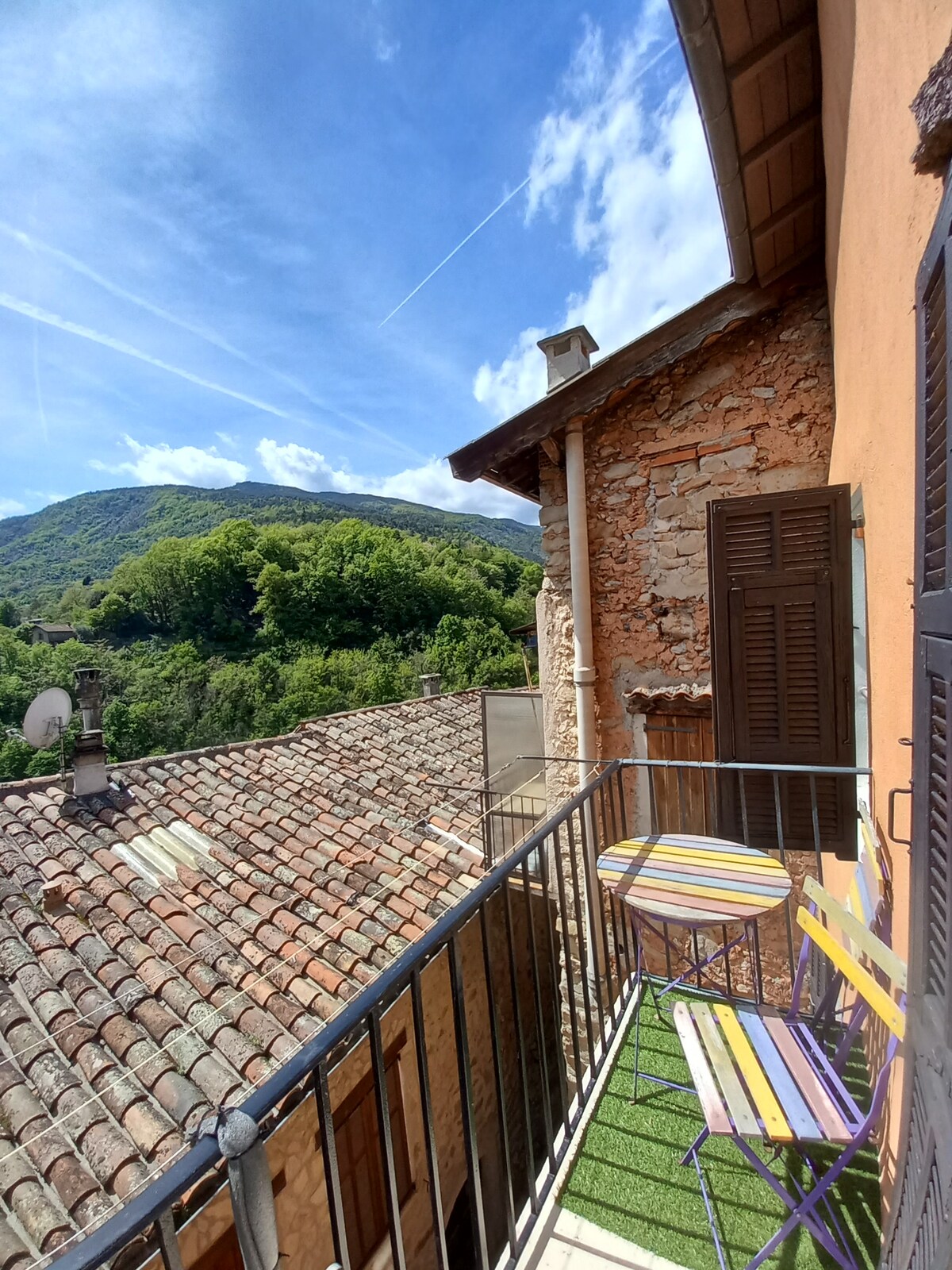 A balcony is visible, featuring two striped chairs positioned on a green patch of artificial grass. The view overlooks terracotta rooftops and lush, green hills under a clear blue sky adorned with white clouds.