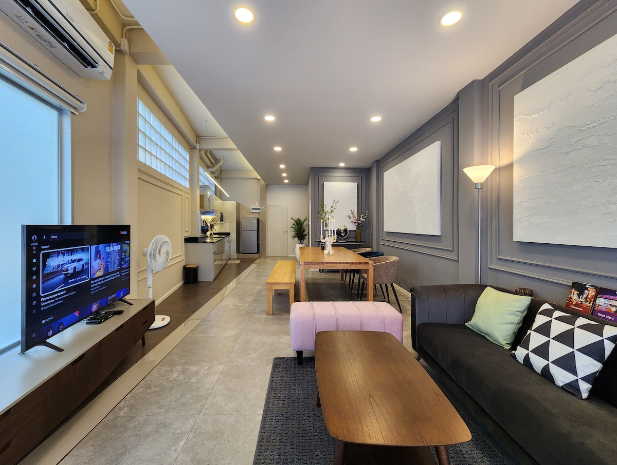 The open living and dining area features a modern design with a large table suited for meals or work. A cozy seating arrangement includes a sectional sofa and coffee table. Natural light fills the space through wide windows, complemented by ambient lighting throughout.