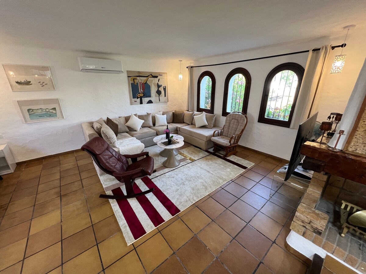 A cozy living area features a spacious sectional sofa adorned with neutral pillows, complemented by a rustic coffee table. Arched windows allow natural light to fill the room, while a rocking chair and decorative wall art enhance the inviting setting.