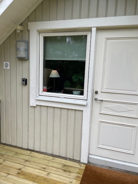 The entrance to the apartment features a white door with a simple design, alongside a large window adorned with a neutral curtain. A light is mounted beside the door, and wooden decking provides access to the entry.