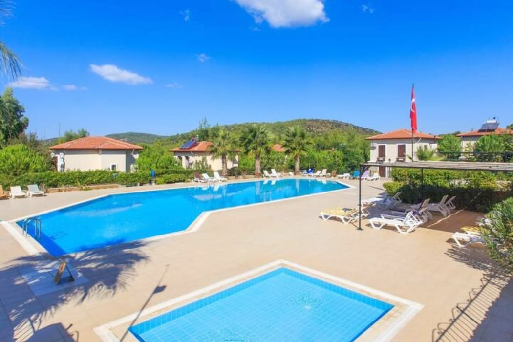 A spacious outdoor area features a large swimming pool surrounded by green foliage and sun loungers. A smaller children's pool is visible nearby. The bright blue sky and distant hills create a serene backdrop, complemented by well-maintained villas along the perimeter.