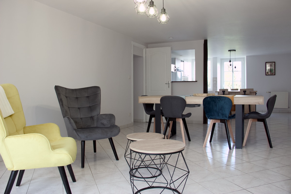 A light-filled living area features a wooden dining table accompanied by several chairs in various colors. Two patterned armchairs are positioned nearby, alongside a modern coffee table with a geometric design. The clean, white walls and tiled floor create a spacious ambiance.