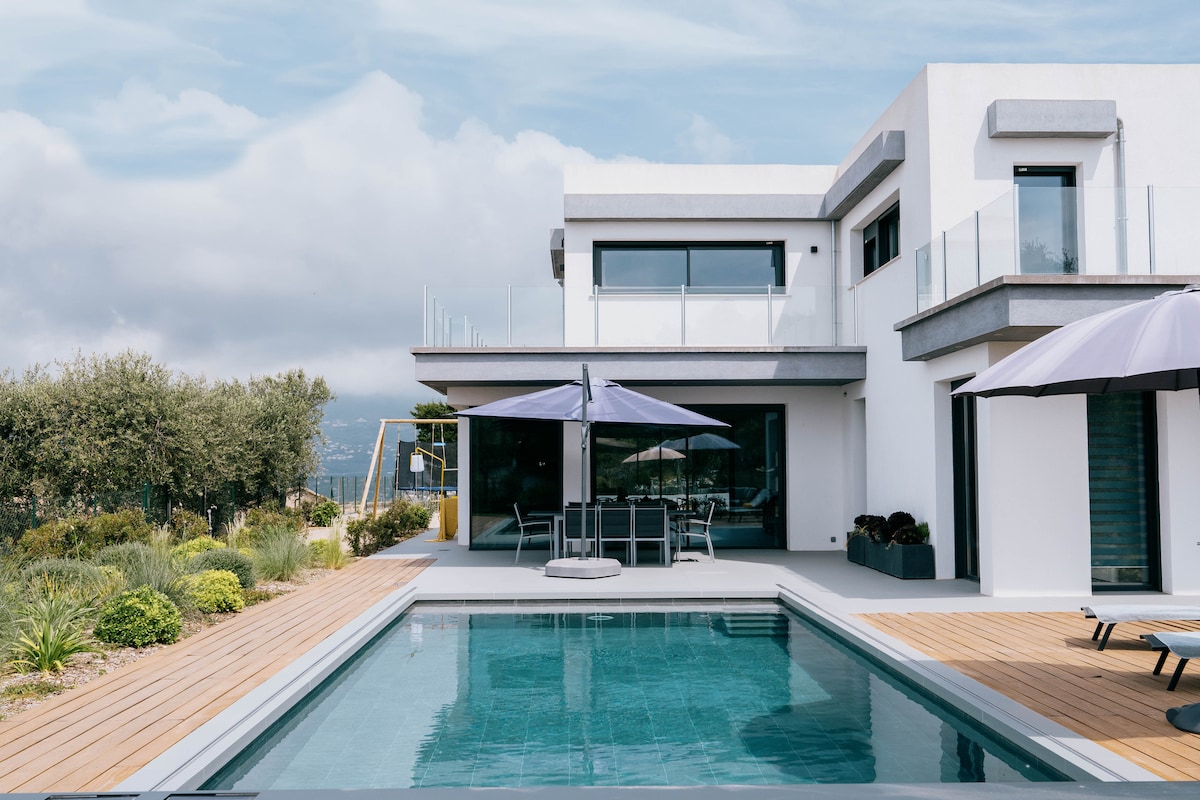 The villa's exterior features a modern design with large windows and multiple outdoor seating areas. A rectangular pool is bordered by wooden decking, complemented by sun umbrellas. Lush greenery and landscaped gardens enhance the tranquil setting, inviting relaxation and enjoyment of the scenic views.