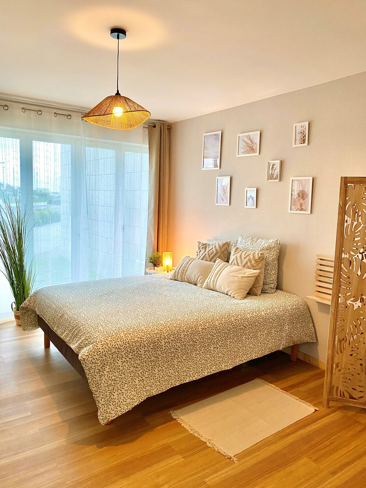 A calming bedroom features a spacious bed dressed in a patterned coverlet, surrounded by decorative pillows. Natural light filters through sheer curtains, illuminating the room. A rattan light fixture hangs above, contributing to a serene atmosphere, while framed botanical art adorns the wall.