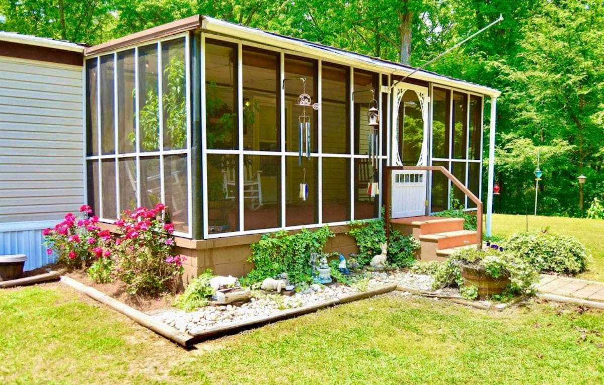 A screened-in porch is framed by large windows, allowing natural light to fill the space. Planters with vibrant flowers are positioned near the entrance, and a small staircase leads down to a grassy area. Decorative stones outline the porch, enhancing the outdoor ambiance.
