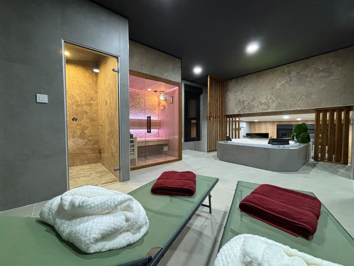 A wellness area features a sauna and steam room, with soft lighting enhancing the space. Two lounge chairs are positioned on a tiled floor, accompanied by rolled white towels and deep red bath linens. The neutral-toned walls and modern design create a relaxing environment.