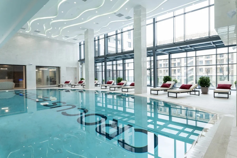 An indoor swimming pool is featured, surrounded by lounge chairs with red cushions. Large glass windows provide natural light and views of the exterior greenery. Sleek, modern design elements are evident in the soft ceiling lighting and elegantly tiled flooring.