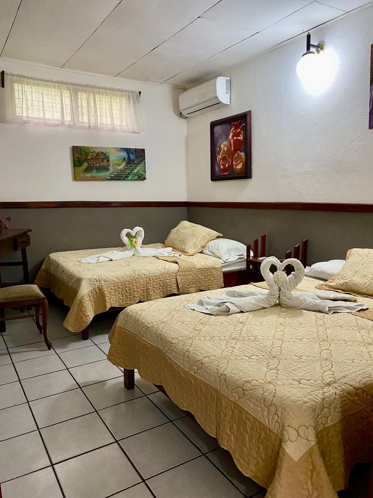 A cozy sleeping area is presented with two spacious beds, each adorned with light yellow quilts. Decorative towels shaped like swans are positioned at the foot of both beds. Natural light filters through a small window, and wall art adds a touch of color.