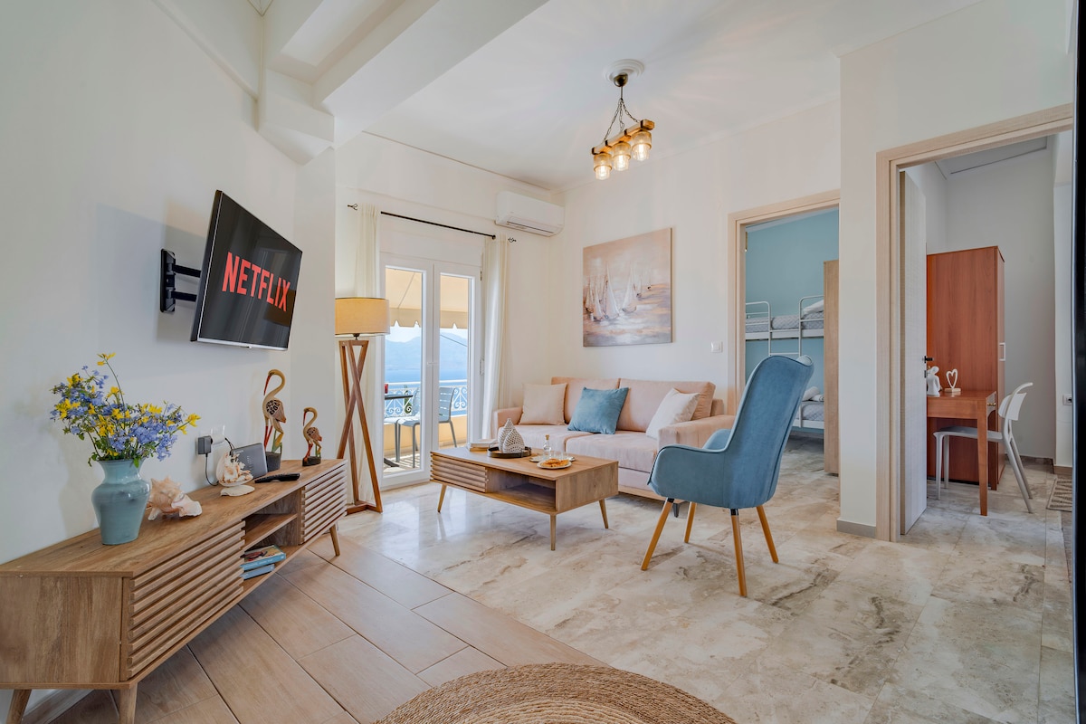 A bright and airy living area features a comfortable sofa and a light blue accent chair. A wooden coffee table displays decorative items. A 50" smart television is mounted on the wall, while natural light floods the space through the window leading to the balcony.