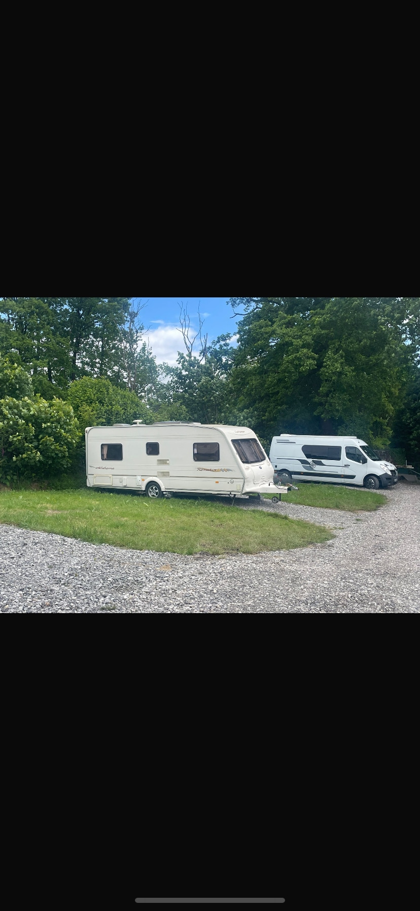 A white caravan is positioned on a gravel area, surrounded by lush greenery. A nearby vehicle is parked adjacent to the caravan. The sky above is clear, suggesting a pleasant day.