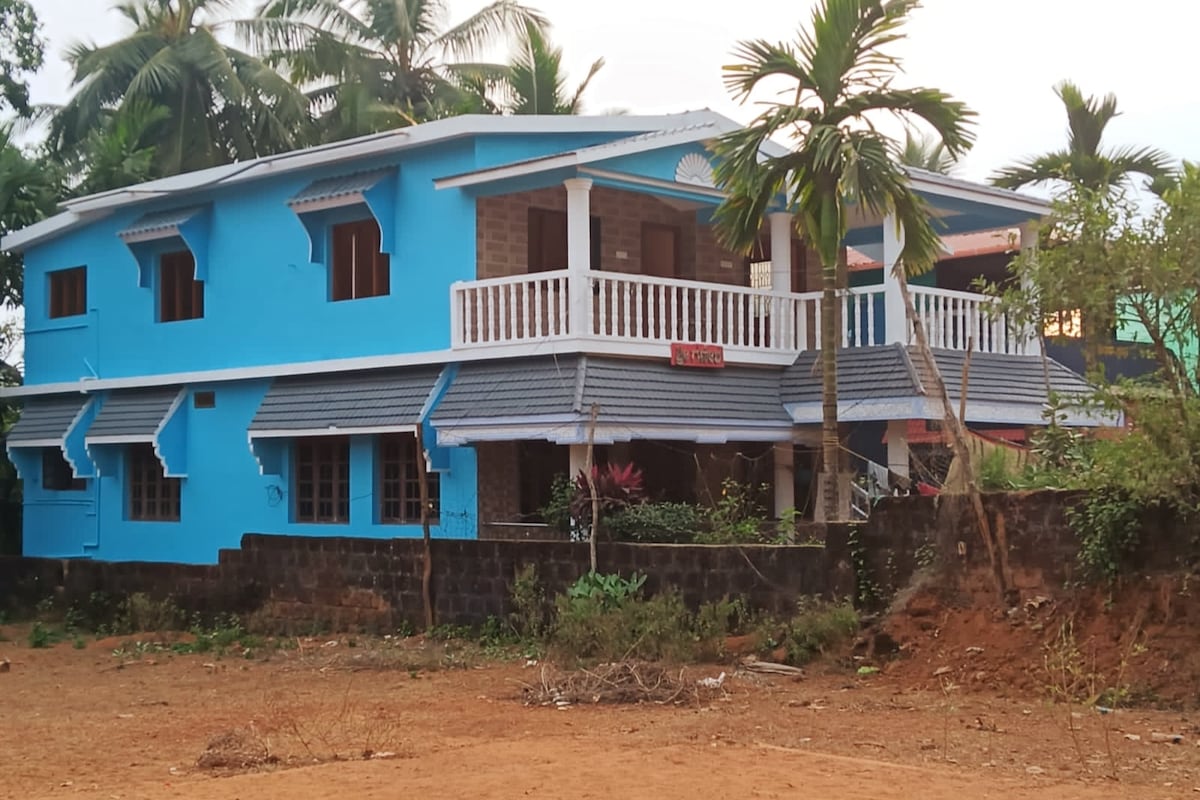 An exterior view of a two-story blue house is shown, featuring a balcony with white railings. Windows are arranged symmetrically along the facade, with tropical plants visible in the surrounding area. The property is set against a natural landscape, with a dirt path and greenery in the foreground.