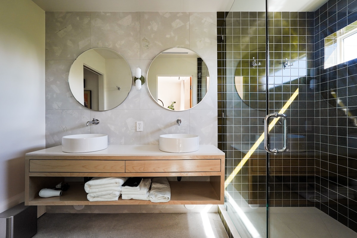 A modern bathroom is presented, featuring dual circular mirrors above a wooden vanity with two vessel sinks. The space is complemented by a glass-enclosed shower showcasing a tiled wall with a subtle color gradient. Fresh white towels are neatly stored beneath the vanity.