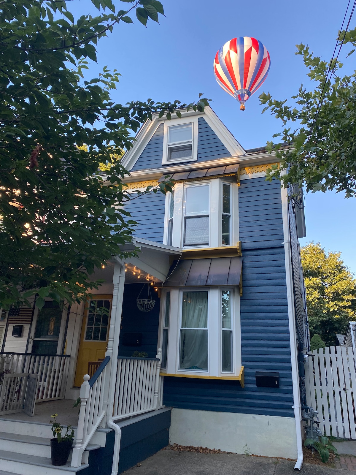 A charming blue duplex with yellow accents stands framed by trees and greenery. Large windows allow natural light to enter. Above, a colorful hot air balloon hovers in a clear blue sky, adding a whimsical touch to the scene.