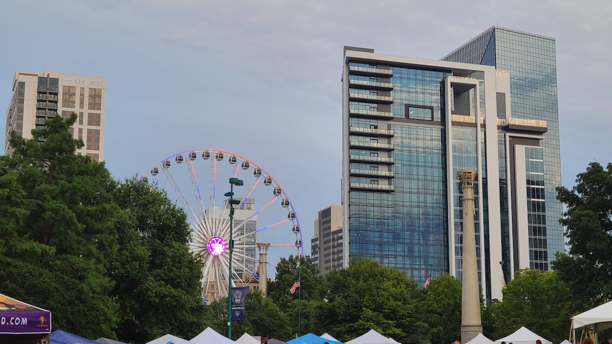 A modern skyline is depicted, featuring tall glass buildings on either side. A large Ferris wheel is visible among green trees, adding a vibrant element to the urban landscape. Tents are set up in the foreground, contributing to a lively atmosphere.