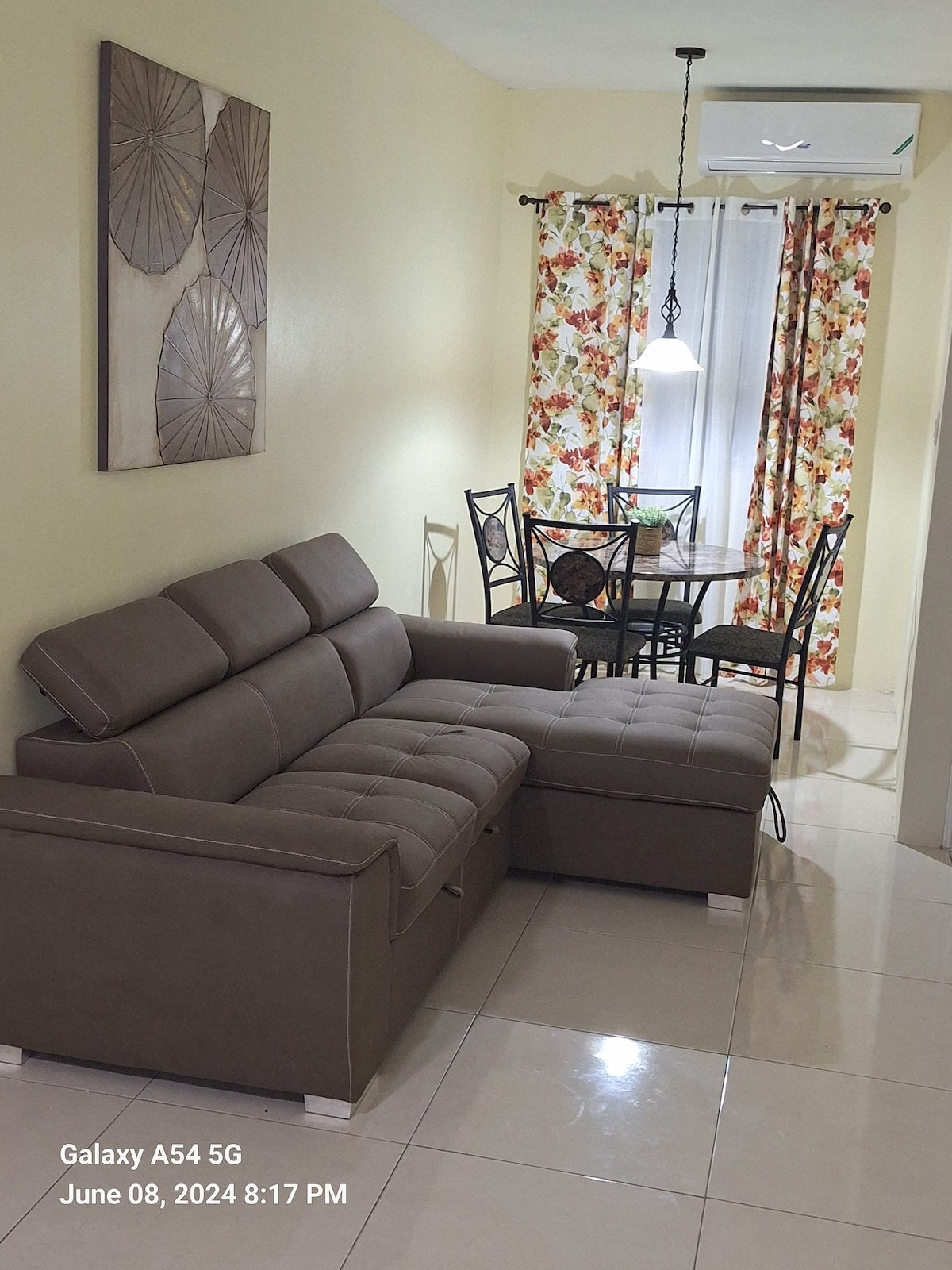 A modern living area features a comfortable sectional sofa in neutral tones, positioned adjacent to a small dining table with intricate metal chairs. Bright floral curtains frame a window, while a pendant light provides illumination in the space.