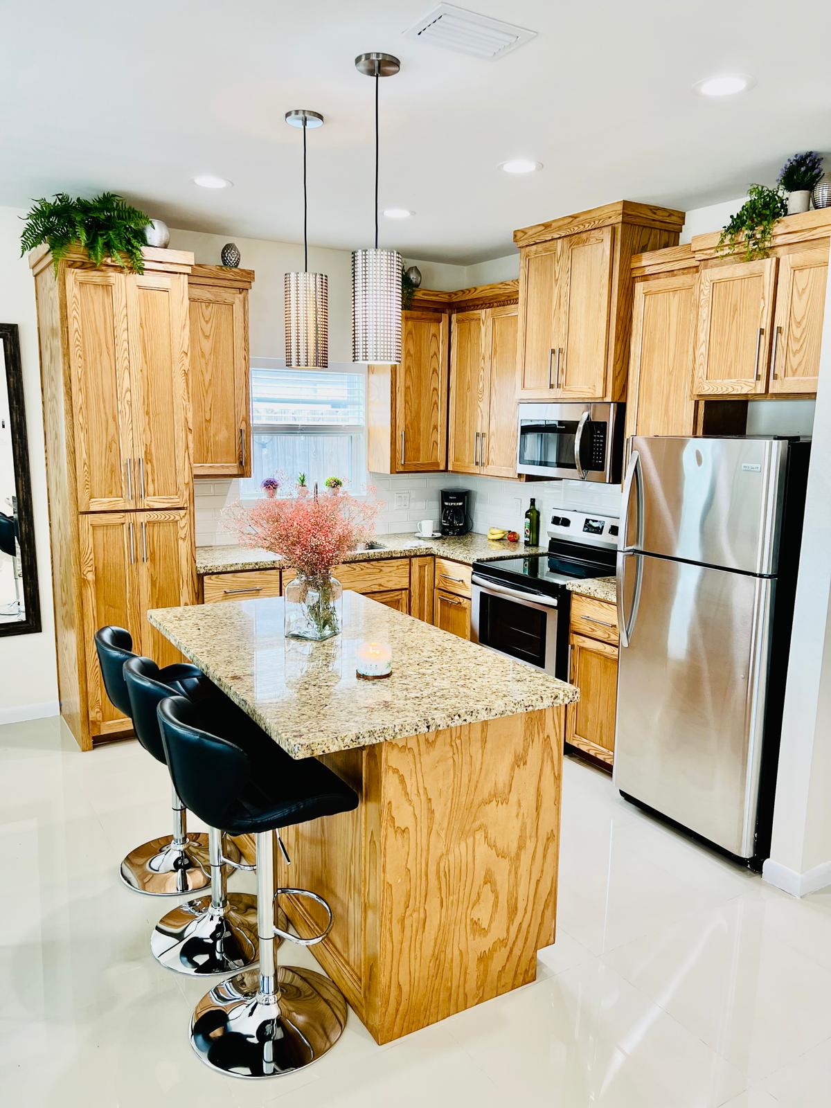 The kitchen features wooden cabinetry complemented by a granite countertop and modern stainless steel appliances. An island with three barstools provides seating. Natural light enters through the window, while pendant lights add soft illumination, enhancing the functional and inviting design.
