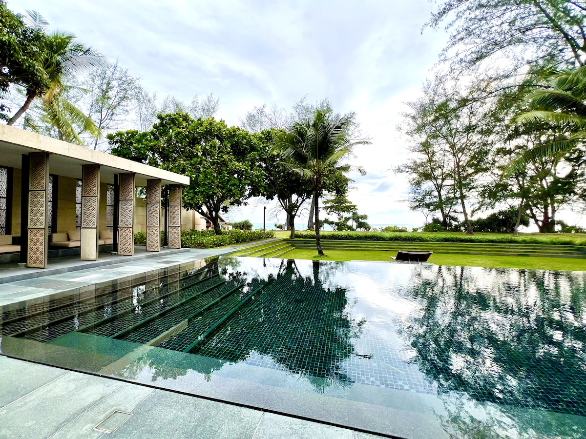 An infinity pool mirrors the sky, surrounded by lush greenery and palm trees. The area features marble tiles and a serene atmosphere, offering a relaxing space to unwind while enjoying the landscape view that extends towards the beach.