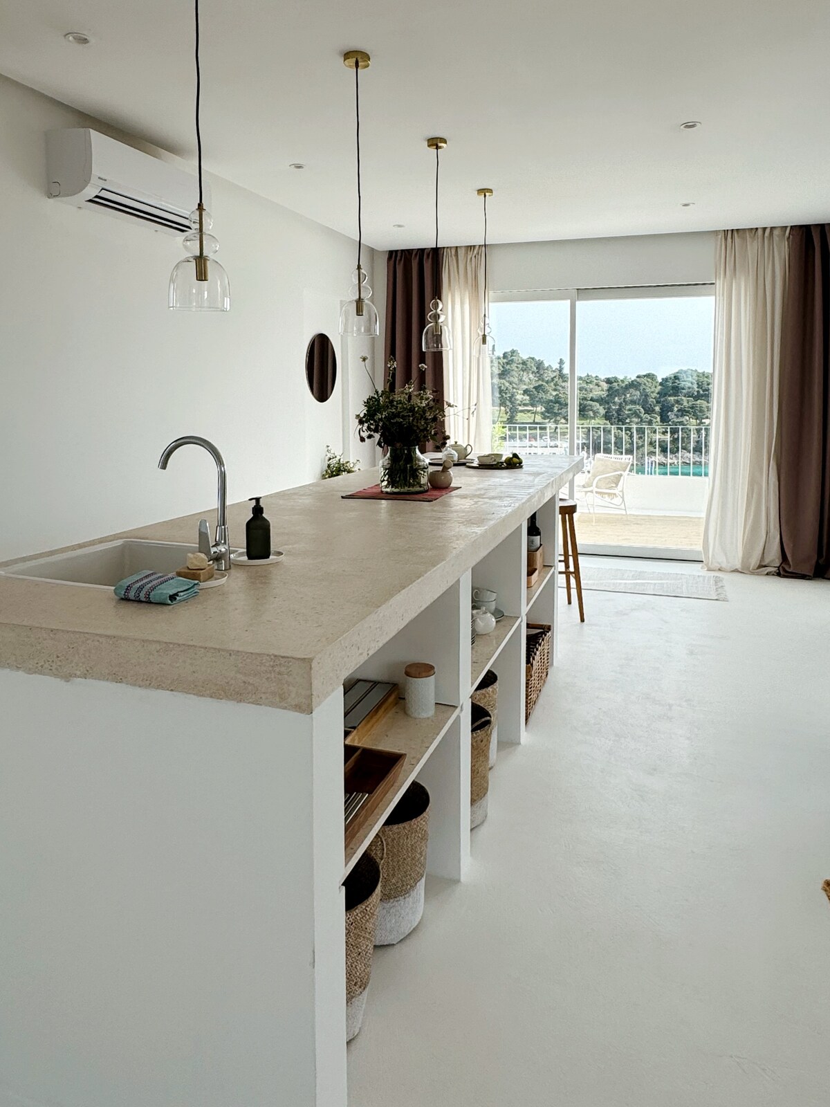 A sleek kitchen area features a long concrete counter, enhancing usability. Transparent pendant lights provide illumination above. Open shelving displays various kitchen essentials, while a window offers a view of the surrounding greenery. A small dining space includes wooden stools for casual seating.