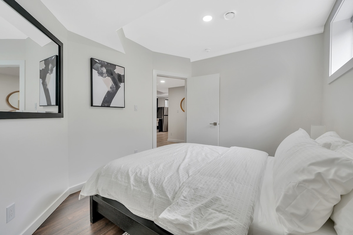 A bright bedroom features a spacious bed with neatly arranged white linens. A large mirror reflects the room, enhancing the sense of space. Soft natural light enters through a window, while the neutral color palette creates a calming atmosphere.