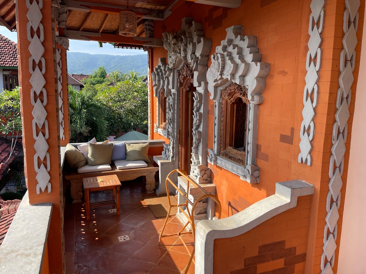 A private outdoor seating area features a comfortable couch and a wooden coffee table set upon terracotta tiles. Intricate carvings adorn the walls, complemented by natural light filtering through lush greenery, creating a tranquil environment with mountain views in the background.