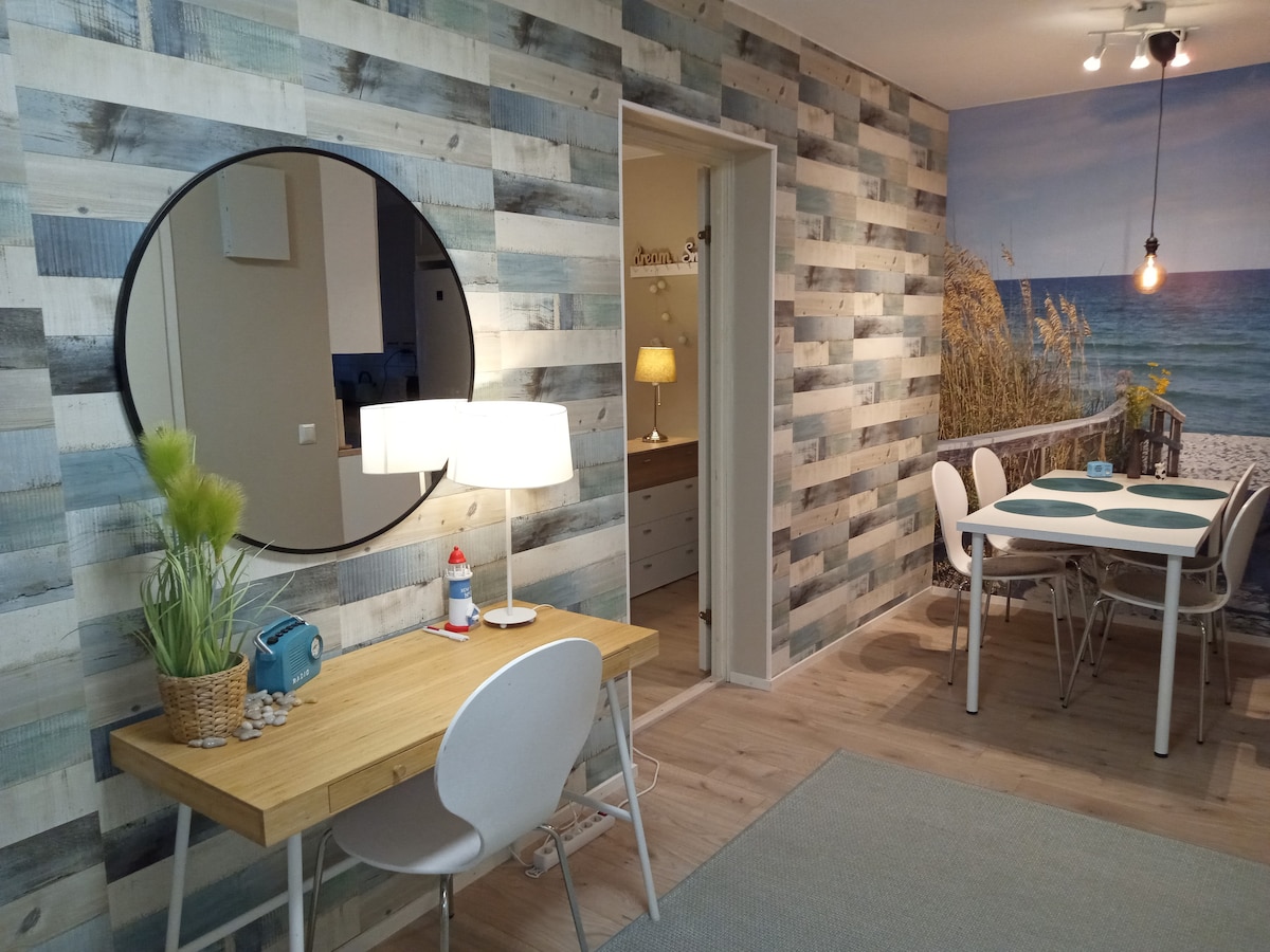 A dining area is featured with a light-colored table and white chairs, set against a beach-themed wall mural. A round mirror is mounted above a wooden desk with a lamp, while greenery adds a natural touch. Soft lighting enhances the welcoming feel.