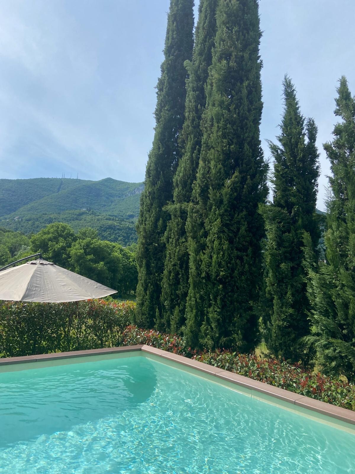 A clear view of a tranquil pool, surrounded by lush greenery and tall cypress trees. A sun umbrella provides shade nearby, with hills visible in the background, creating a serene outdoor space for relaxation.