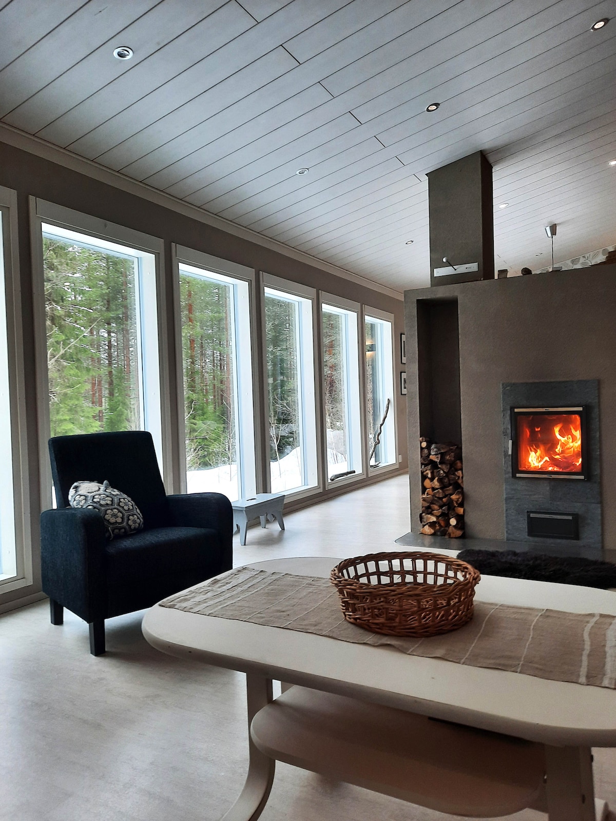 A comfortable seating area is highlighted by a dark armchair positioned beside large windows, providing views of the surrounding trees. A modern fireplace is visible, with logs stacked nearby. A simple dining table with a textured runner and a woven basket sits in the foreground.