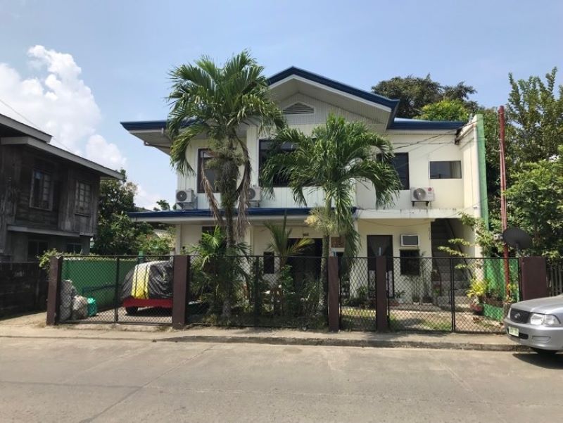 A two-story building is framed by lush greenery and palm trees, featuring a light exterior with blue accents. Air conditioning units are mounted on the walls, and a fenced yard is visible in front, creating a welcoming entryway.